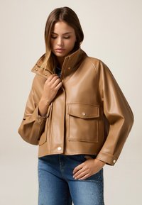 Giacca in ecopelle marrone con colletto alto, chiusura con zip e due tasche frontali. Presenta maniche over e una texture liscia e lucida.