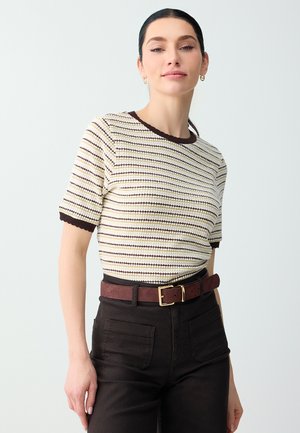 Vrouw draagt een kort mouw, gestreepte gebreide top met donkere rand, gecombineerd met zwarte broeken met hoge taille en een bruine riem met gouden gesp.