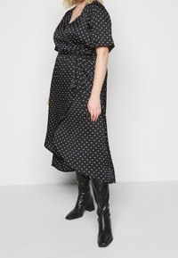 Schwarzes Wickelkleid mit Po-Lka Punkten, kurzen Ärmeln, einem V-Ausschnitt und einem asymmetrischen Saum, kombiniert mit knielangen schwarzen Stiefeletten mit hohen Absätzen.