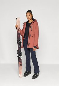 Smučarska oprema z vodoodporno jopico rjave barve, črnim puloverjem, mornarsko modrimi hlačami in črnimi smučarskimi čevlji. Drži svetlo roza smučko s vezi.