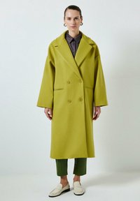 Manteau double boutonnage vert lime avec large col, coupe oversize et grands boutons, associé à un pantalon cropped vert foncé et des mocassins.