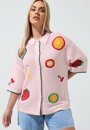 Kvinde iført en pink strikket cardigan med farverige frugt mønstre og lyseblå denimshorts, stående mod en enkel lys baggrund.