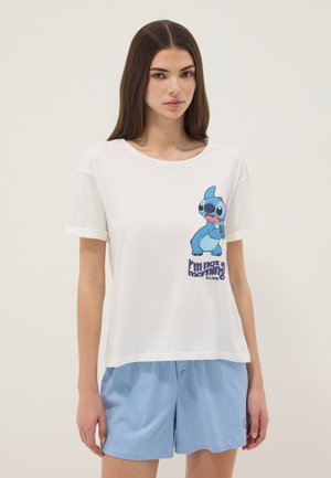 Jonge vrouw draagt een wit T-shirt met een blauwe cartoon alien en tekst, gecombineerd met lichtblauwe shorts, staand tegen een effen achtergrond.