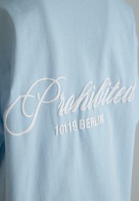 Light blue cotton t-shirt featuring white embroidered text "Prohibited" and "10119 Berlin" on the back, with a soft texture.