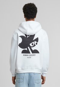 Sweat à capuche blanc avec un grand design graphique noir présentant des formes abstraites et des yeux au dos. Le tissu semble doux et texturé.