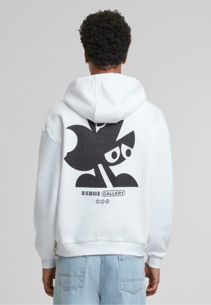 Hvit hettegenser med stort svart grafisk design som viser abstrakte former og øyne på baksiden. Stoffet ser mykt og teksturert ut.