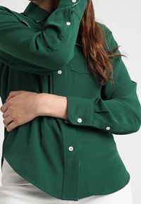 Camicia verde a maniche lunghe con chiusura a bottoni, tasca e cuciture bianche a contrasto. Tessuto liscio con vestibilità rilassata e orlo curvo.