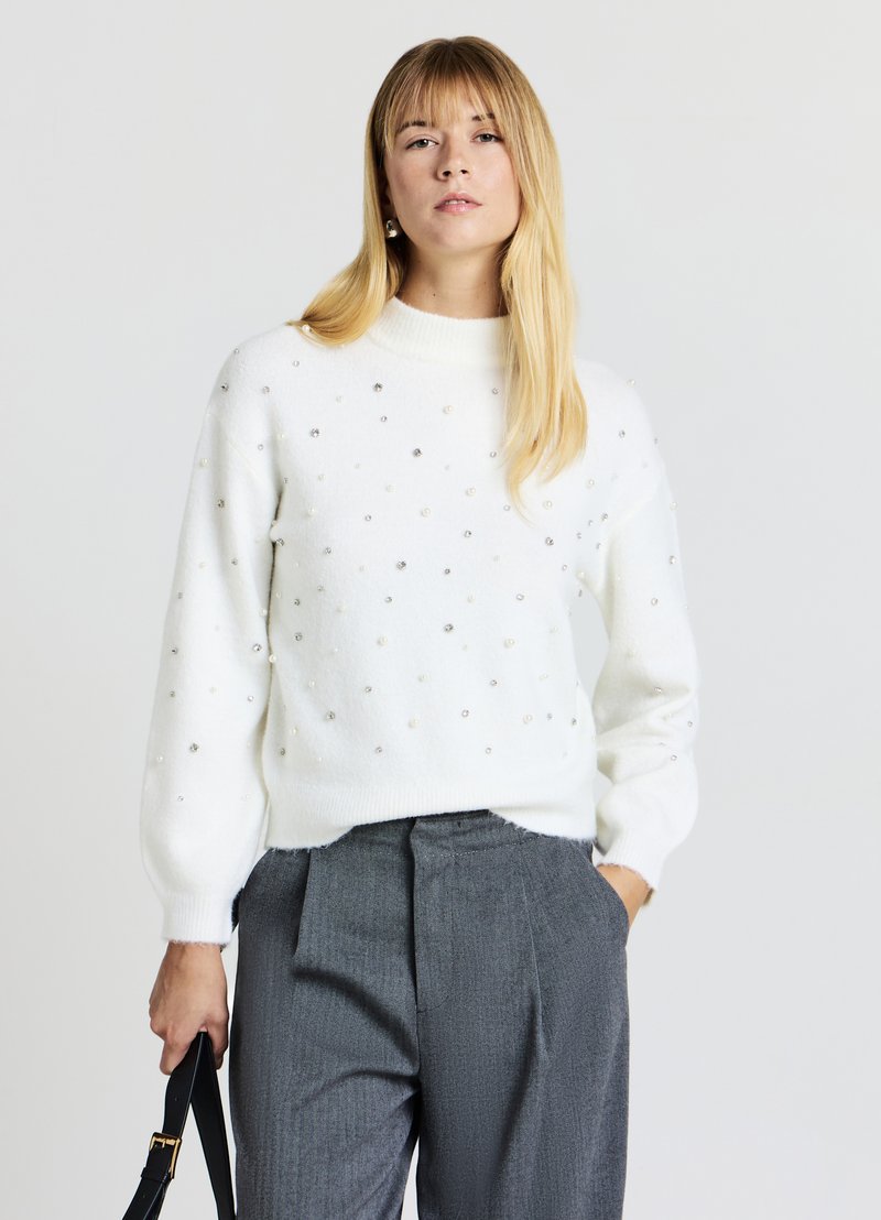 Pull en tricot blanc avec un col rond et une surface texturée. Embelli de petits accents argentés et ressemblant à des perles. Pantalon gris ajusté.