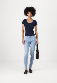 Mørkeblå v-hals t-skjorte, tettsittende design. Lys blå skinny jeans, lett slitasje. Svarte flate sko. Svart veske med skulderrem.