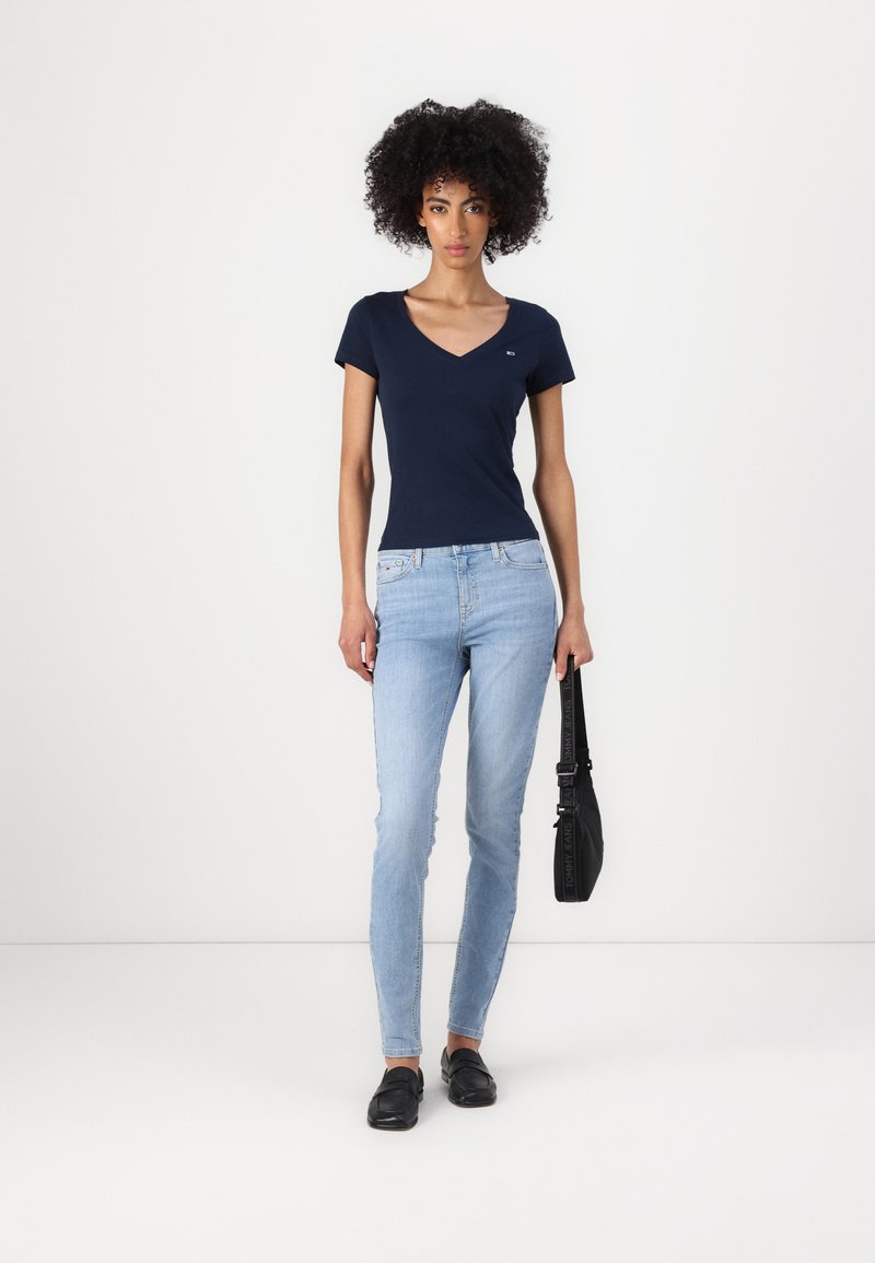 Mørkeblå v-hals t-skjorte, tettsittende design. Lys blå skinny jeans, lett slitasje. Svarte flate sko. Svart veske med skulderrem.