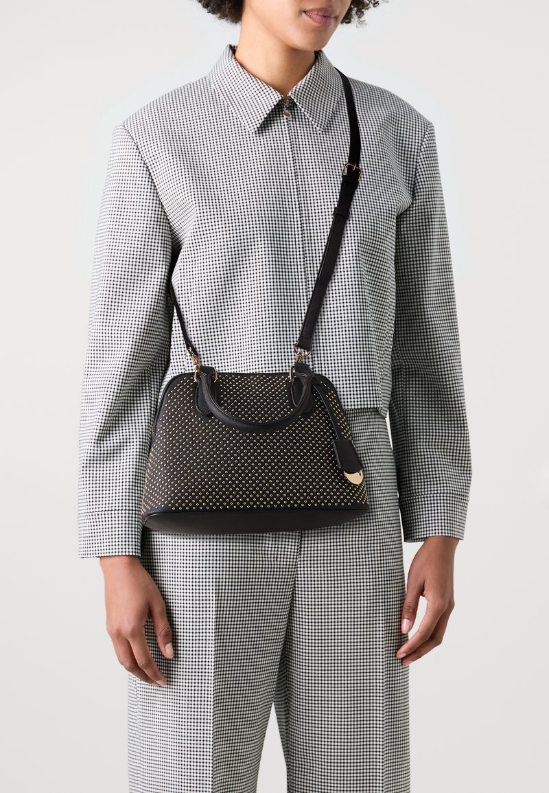 Femme portant un tailleur à carreaux noir et blanc tenant un sac à main noir avec des clous dorés et une bandoulière noire en travers de la poitrine.