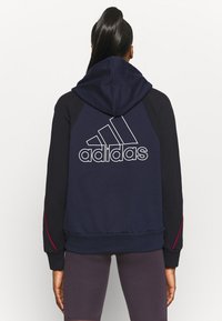 Persona con cabello largo recogido en una cola de caballo alta, vestida con una sudadera navy y negra de Adidas con el logo en la parte trasera y pantalones cortos ajustados oscuros.