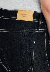 Mørkeblå jeans med synlig merkeetikett "perfect jeans gina tricot" og hvite sømdetaljer nær en lys grå topp.
