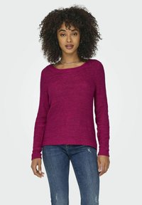 Pull en tricot fuchsia avec un large col, tissu texturé et manches longues, associé à un jean bleu avec des détails déchirés.