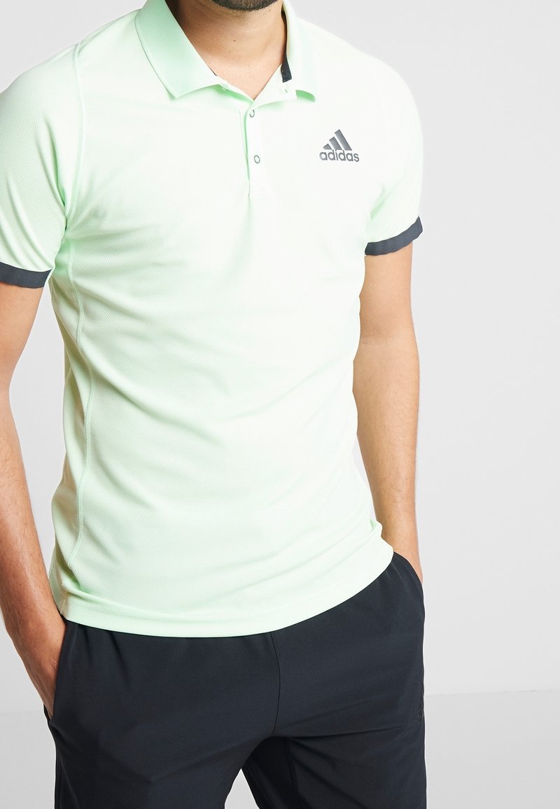 Polo shirt vert clair avec un tissu texturé, col et poignets noirs, et un logo Adidas sur la poitrine gauche. Associé à un pantalon sombre.
