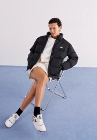 Svart pufferjakke med glidelås og logo på brystet, kombinert med en hvit kjole og hvite sneakers, sittende på en stol mot en blå bakgrunn.