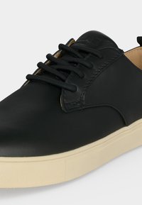 Basket en cuir noir avec des lacets noirs et une épaisse semelle en caoutchouc blanc sur fond gris.
