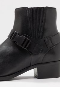 Bottine en cuir noir avec un petit talon carré et une sangle ajustable avec une boucle en plastique autour de la cheville.