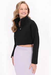 Jachetă neagră, scurtă, cu guler înalt și închidere cu fermoar pe jumătate. Textură netedă și mâneci lungi, asortată cu o fustă violet deschis.