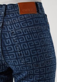 Modré džíny z denimu s texturovaným geometrickým vzorem. Mají hnědou koženou pásku s embosovaným nápisem "ONLY" a viditelným detailem zadní kapsy.