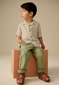 Lichtbeige korte mouwen overhemd met knopen, groene broek met opgerolde zomen en bruine sandalen. Kind zittend op een pastelroze blok. Neutrale achtergrond.