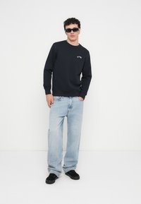 Fekete pulóver bordázott mandzsettákkal és gallérral, világoskék bő szárú farmernadrág és fekete sportcipő. A modell sötét napszemüveget visel, fehér háttér előtt áll.