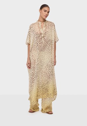 Femme portant une tunique ample crème et un pantalon large assorti à pois marron, debout sur un fond blanc uni.