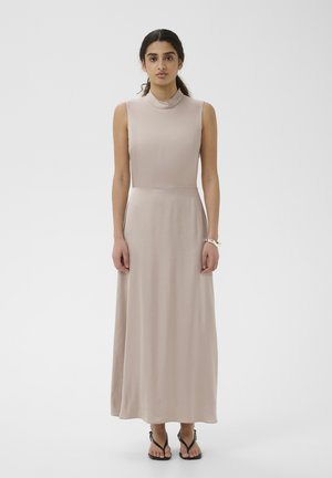 Kvinde med mørkt hår i en lav hestehale iført en ærmeløs beige maxikjole og sorte sandaler, stående mod en ensartet hvid baggrund.