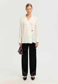 Weißes langärmliges Wickelshirt mit V-Ausschnitt und dekorativem Knopf, kombiniert mit schwarzen weit geschnittenen Hosen und einer kleinen lila Handtasche.