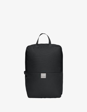 Sac à dos en tissu noir de forme rectangulaire avec poignée supérieure, boucle latérale et petit écusson logo sur le devant, présenté sur un fond blanc.