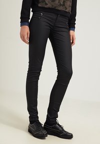 Černé skinny kalhoty vyrobené z hladkého elastického materiálu, s kovovými akcenty a zipy, doplněné černými botami s cvočky.