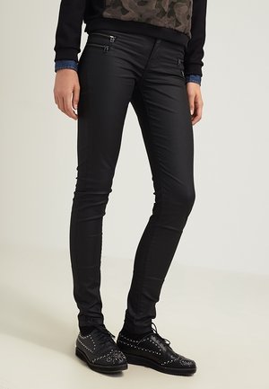 Mustat tiukat housut, jotka on valmistettu sileästä, venyvästä kankaasta, metallikoristeilla ja Vetoketjuilla, yhdistettynä mustiin niittikenkäisiin.