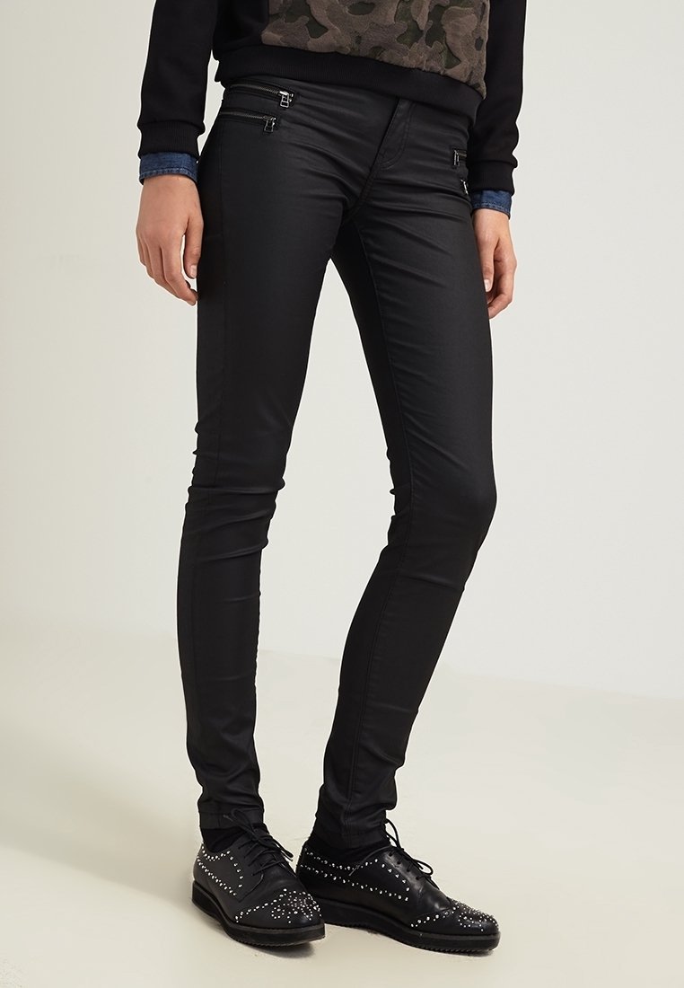 Černé skinny kalhoty vyrobené z hladkého elastického materiálu, s kovovými akcenty a zipy, doplněné černými botami s cvočky.