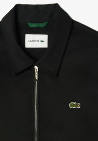 Melnā jakā ar apkakli, kas aprīkota ar priekšējo rāvējslēdzēju, sieta oderi un mazu zaļu krokodila logotipu izšūtu uz krūšu.