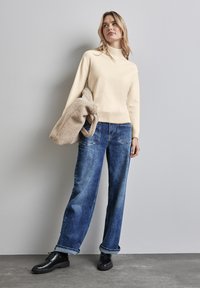 Maglione beige lavorato a maglia con collo alto, abbinato a jeans di denim blu larghi. Scarpe nere con lacci e una borsa beige strutturata completano il look.