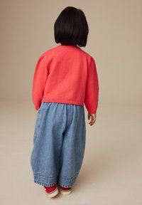 Pull en tricot rouge à manches longues, pantalon en denim bleu large et baskets rouges. La vue arrière montre des manches bouffantes et des jambes de pantalon larges, avec des poignets côtelés.