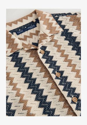 Rövid ujjú ing, amely krém, barna és tengerészkék zigzag mintával rendelkezik, gombos gallérral és fa gombokkal. Strukturált szövet.