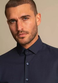 Lühikeste tumedate juuste ja habemega mees, kes kannab tumedat sinist nööbitavat särki, vaatab otse ette neutraalse ilmega.