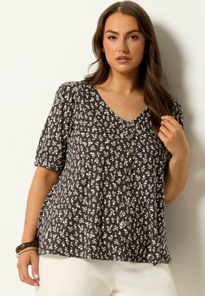 Kvinde iført en sort knap-op bluse med hvidt blomsterprint og hvide bukser, poserer med en hånd nær håret og armbånd på håndleddet.