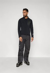 Zwarte jas met ritssluiting en een gestructureerdborstpaneel, gecombineerd met donkergrijze broek. Model draagt zwarte skischoenen met metalen details.