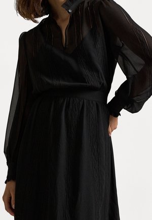 Frau trägt ein schwarzes langärmeliges Kleid mit transparenten Ärmeln und vertikalen Stickmustern, posiert mit einer Hand auf der Hüfte.