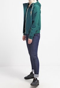 Teal hættejakke med lynlås foran, ribbede manchetter og et lille brunt mærke; båret over mørkeblå jeans og sorte sko.