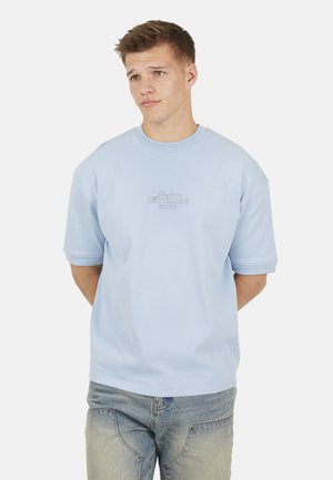 Ragazzo con indosso una camicia a maniche corte azzurro chiaro con la scritta "Amsterdam", in piedi con le mani dietro la schiena, che guarda leggermente di lato.