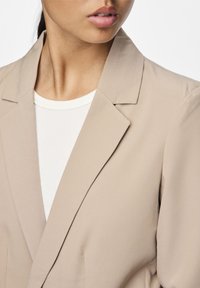 Beige sako s hladkou texturou, s klopami a přiléhavým střihem, nošené přes bílý ribovaný top. Záběr na límec a výstřih.