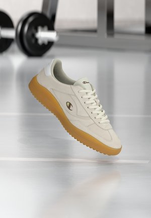 Witte sneaker met gumzool en Champion-logo, zwevend boven de gymvloer, halters op de achtergrond vervaagd.