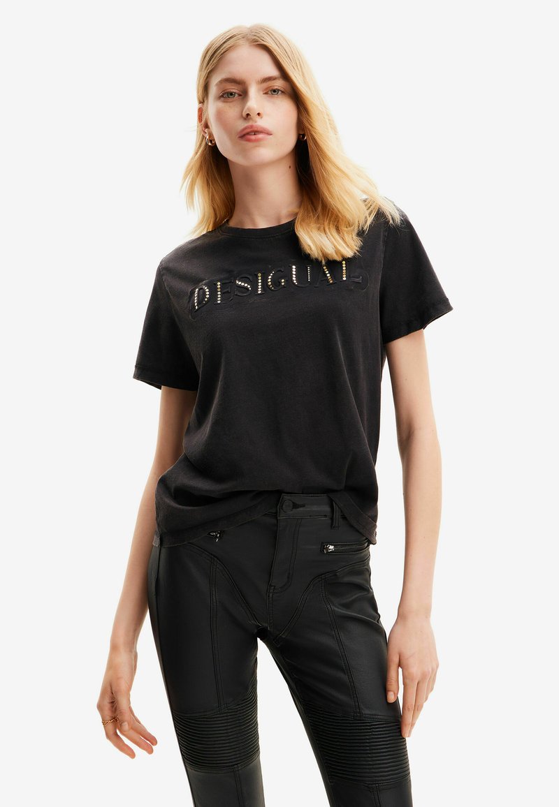 Camiseta de algodón negra con un logo texturizado y acentos metálicos. Manga corta y corte holgado. Combinada con pantalones negros con cremallera.