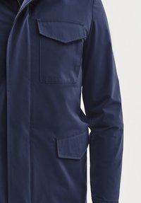 Granatowa wodoodporna kurtka z wysokim kołnierzem, dwoma przednimi kieszeniami i zapięciem na zamek, o gładkiej fakturze i minimalistycznym wzornictwie.