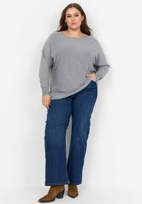 Grauer übergroßer Pullover mit langen Ärmeln, kombiniert mit geraden, dunkelblauen Jeans und braunen, hohen Stiefeletten. Glatte, weiche Stofftextur.