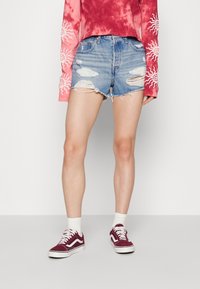 Světle modré džínsové šortky s prošoupáním a třepícími se okraji, v kombinaci s vínovými teniskami Vans a bílými kotníkovými ponožkami. Růžové tričko s dlouhým rukávem a batikováním.