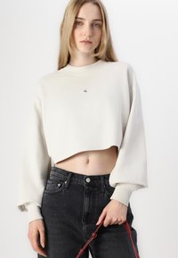 Cropped, beige sweatshirt with long sleeves and ribbed collar. Displayed with high-waisted, dark denim jeans and a red strap purse.